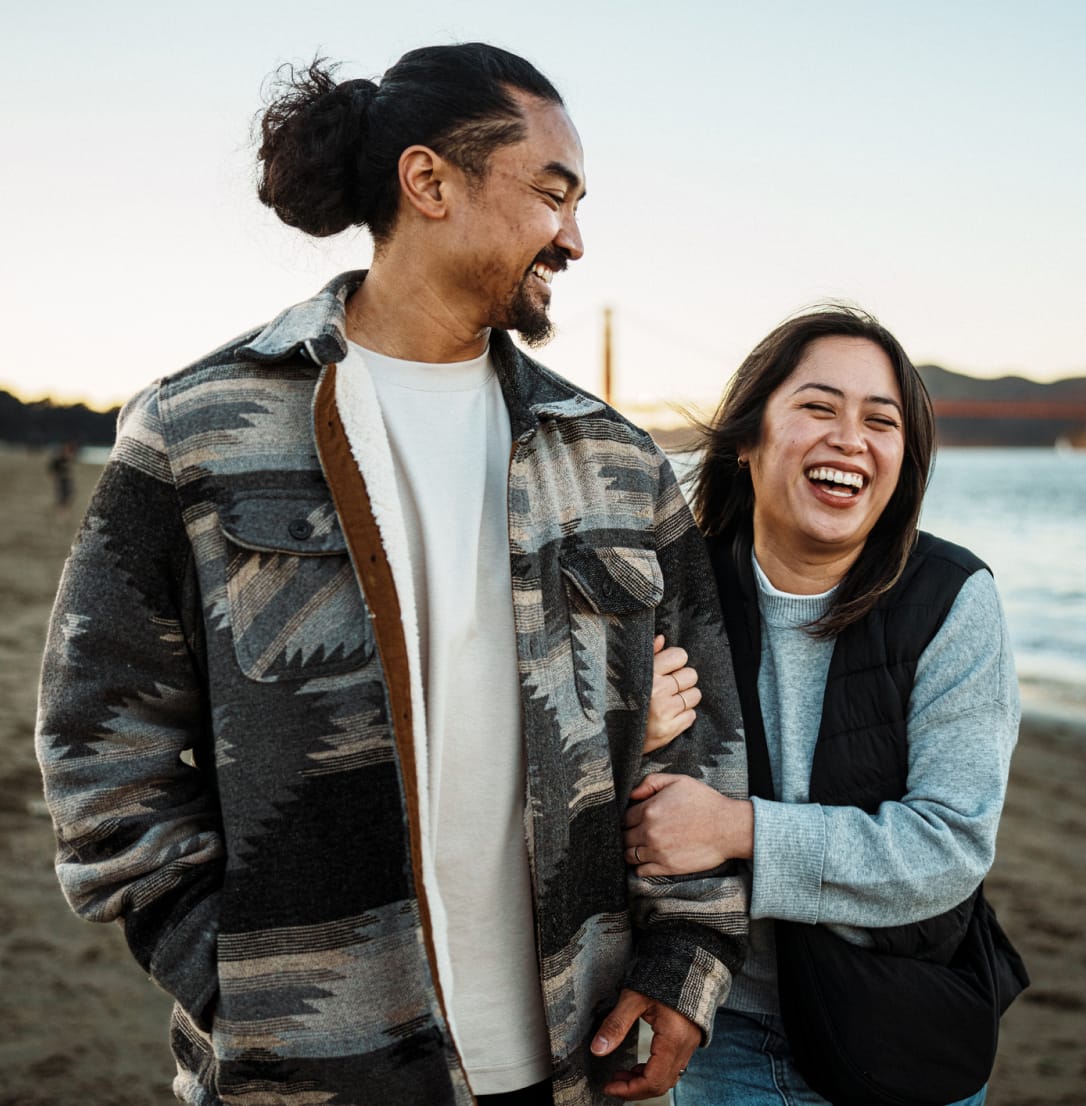 Man and woman laugh with one another as they walk along the beach with view of Golden Gate Bridge in the background.