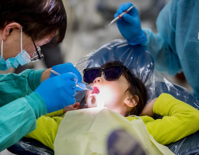 A dentist in protective gear examines a young child's teeth while an assistant holds a tool.