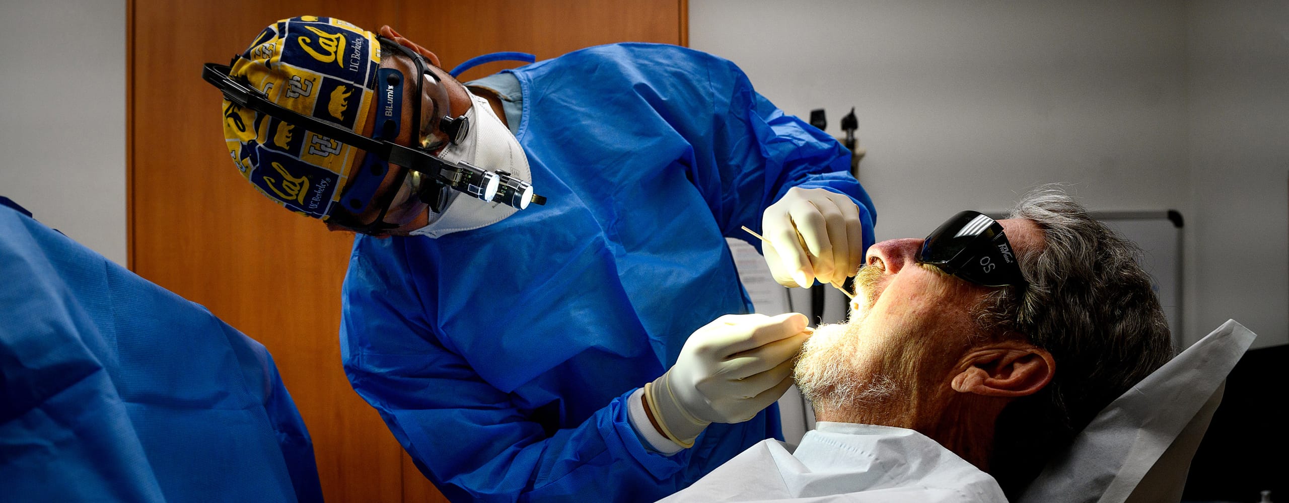 A dental student or alumnus shows school spirit with a UC Berkeley 'Cal' scrub cap while performing a precise clinical examinati