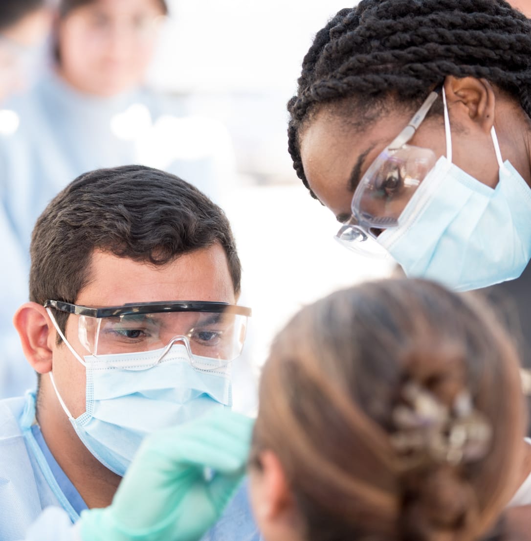 Two healthcare professionals in masks and safety glasses perform a clinical procedure on a patient.